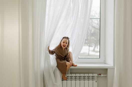 Portrait of a little girl in a brown dressing gown, who is sitting on the windowsill.