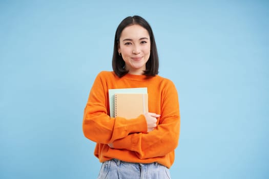 Smiling asian woman with notebooks, student with happy face, promo of college education, blue background.