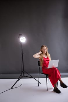 A business woman blogger in a red dress is working, typing on a laptop apple and sitting on a chair, gray background. portrait blonde assistant gaffer of hands with computer MacBook . Vertical