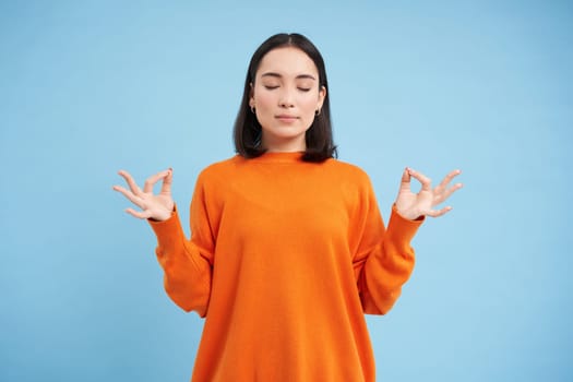 Emotions under control, zen. Young calm asian woman meditating, breathing with relaxed face, standing over blue background.