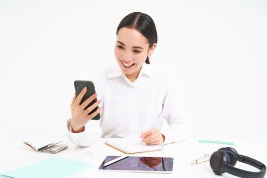 Young woman professional, businesswoman has a meeting on video chat, talks to mobile phone with client, stands over white background.