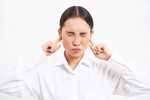 Annoyed young asian corporate worker, lady screams and shuts her ears with fingers, stands frustrated against white studio background.