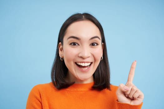 Close up portrait of enthusiastic asian woman, points up and laughs, smiles and looks happy, stands over blue studio background.