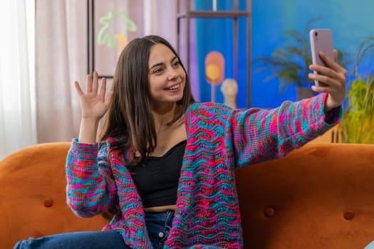 Happy woman blogger taking selfie on smartphone, communicating video call online with subscribers, recording stories for social media. Portrait of Caucasian girl at home apartment living room on sofa