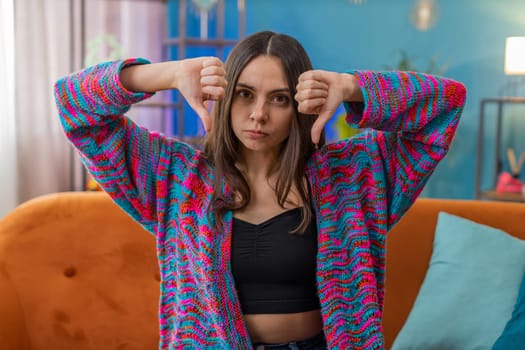 Dislike. Upset young woman showing thumbs down sign gesture, expressing discontent, disapproval dissatisfied bad work at modern home apartment indoors. Displeased Caucasian girl in living room on sofa