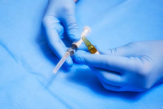 The hands of a nurse in blue protective gloves hold an ampoule and a syringe. Soft filter used on the image