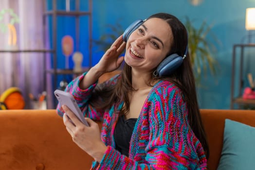 Happy overjoyed Caucasian woman in wireless headphones relaxing sitting on couch at home apartment choosing listening favorite energetic disco dancing music. People weekend daytime leisure activities