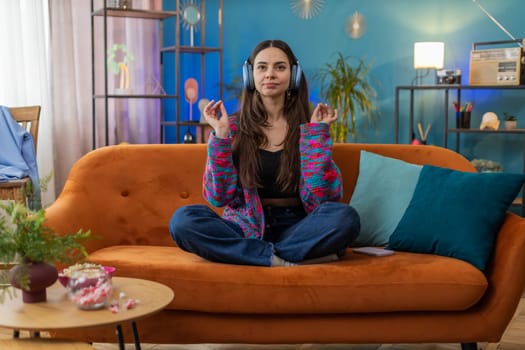 Keep calm down, relax, inner balance. Young woman breathes deeply with mudra gesture, eyes closed meditating with concentrated thoughts, peaceful mind. Portrait of girl at home apartment room on sofa