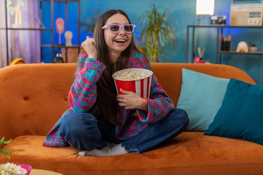 Excited woman in 3D glasses sits on couch eating popcorn snacks and watching interesting TV serial sport game, film online social media movie content at home apartment. Girl enjoying entertainment