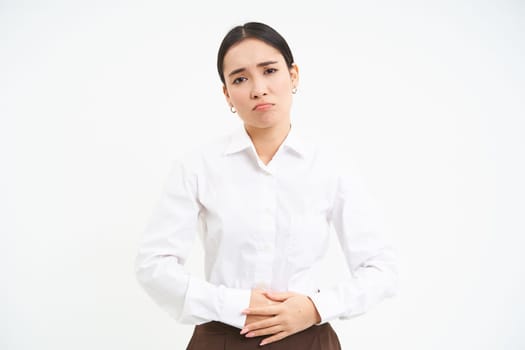 Image of asian office lady touches her belly, looks upset from discomfort in stomach, suffers period cramps at work, white background.