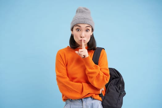 Asian girl shows shush, shh gesture, puts finger over lips, keep quiet sign, stands over blue background with backpack and wears hat.