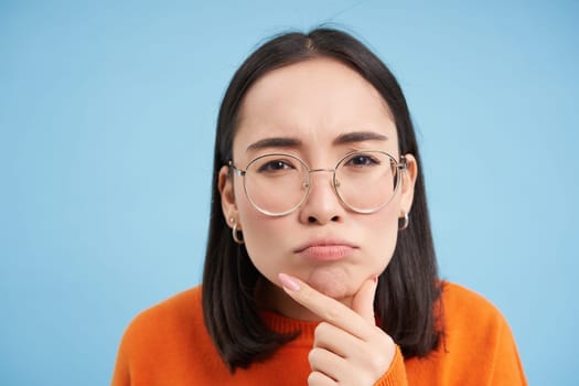 Close up of asian woman in glasses, thinking, staring at camera with thoughtful face expression, standing over blue background.