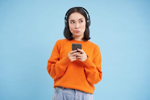 Stylish urban asian girl in headphones, holds mobile phone and looks thoughtful, thinking while choosing music on smartphone app, blue background.