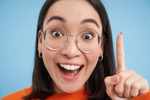Close up portrait of happy asian girl in glasses, raises finger, has revelation, suggest great idea, says eureka, stands over blue background.