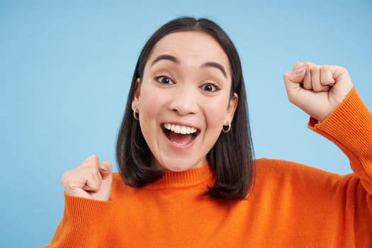 Close up of cheerful japanese woman celebrates, raises hands up and laughs, wins and triumphs, stands over blue background.