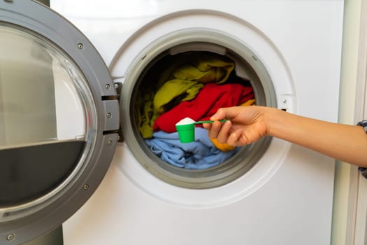 A woman's hand holds a cap with washing powder against the background of a washing machine with bright things put into it. The girl washes and wringes out things in the laundry room at home.