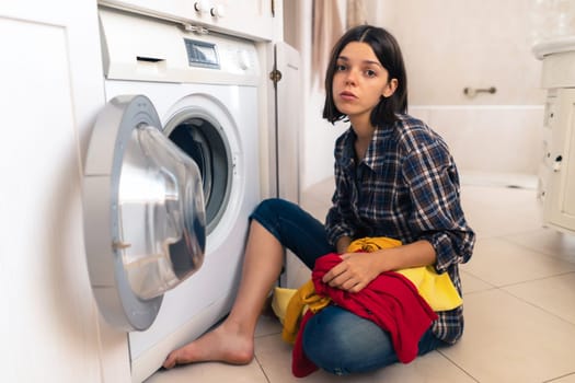 Young girl wearily leaned her elbows on her hand, sits on the bathroom floor next to the washing machine and holds dirty clothes. Woman is tired of household chores, routine and house cleaning.