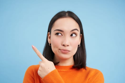 Close up of skeptical asian woman, looks and points left with suspicious face, stands over blue background.