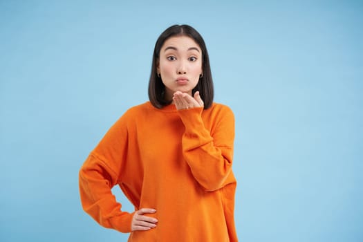 Cute young woman puckers her lips and blows air kiss at camera with coquettish face expression, stands over blue studio background.