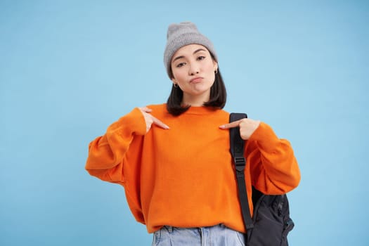 Stylish korean woman points at herself, stands in warm hat and holds backpak on shoulder, blue background. Copy space