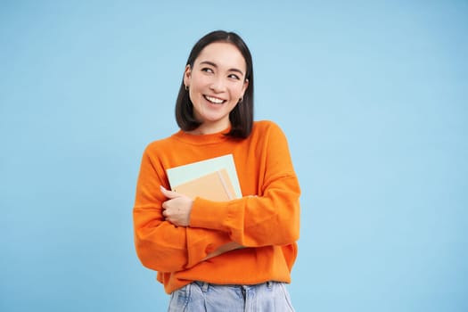 Smiling asian woman with notebooks, student with happy face, promo of college education, blue background.