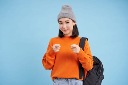 Cute korean woman in hat with backpack, student pointing fingers at camera, inviting you, congratulating, standing over blue background.