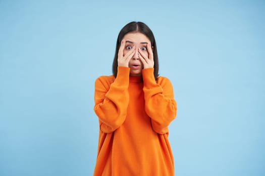 Scared Asian woman shuts her eyes and peeks through hands at smth scary and disturbing, shocked by smth horrifying, standing over blue background.