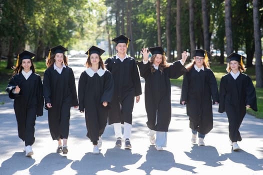 Row of happy young people in graduation gowns outdoors. Students are walking in the park