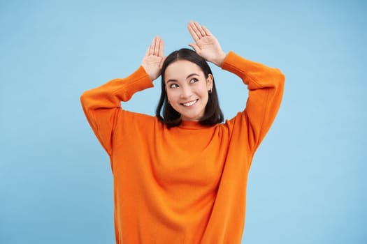 Funny and cute young asian woman shows bunny ears gesture on top of her head and smiling, dancing and showing grimaces, blue background.
