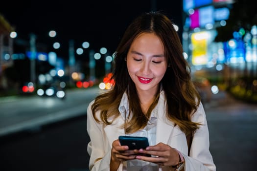 Business woman using mobile phone walking through night city street while waiting car to pick up home, Beautiful young smiling female texting work on smartphone, social media