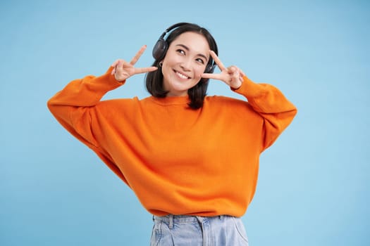 Beautiful korean woman in headphones, dancing and listening music in earphones, standing over blue background.