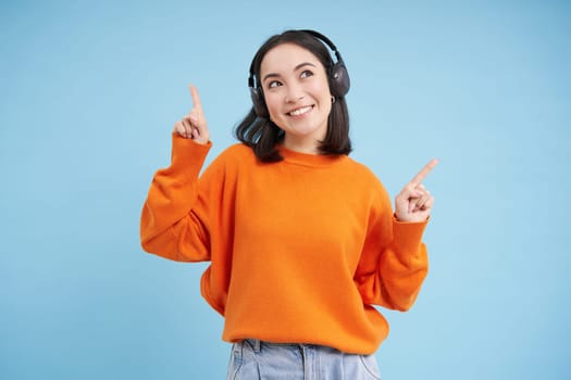 Happy Chinese woman in headphones, listens music, enjoys favourite song in her playlist, stands over blue background. Copy space