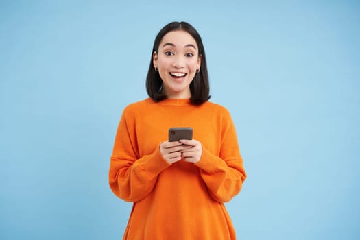 Happy asian woman with smartphone, found great offer online on mobile phone, excited over discounts in app, stands over blue background.