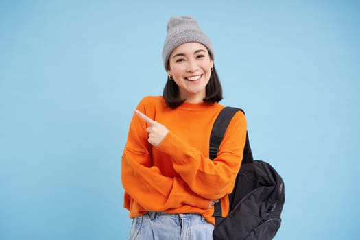 Stylish korean woman in hat, holds backpack, points finger at advertisement, shows promo offer banner, stands over blue background.