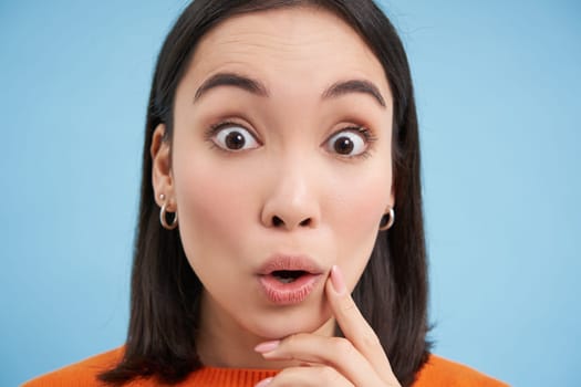 Close up portrait of girl looks with surprised, amazed eyes, says wow, impressed by smth, stands over blue background.