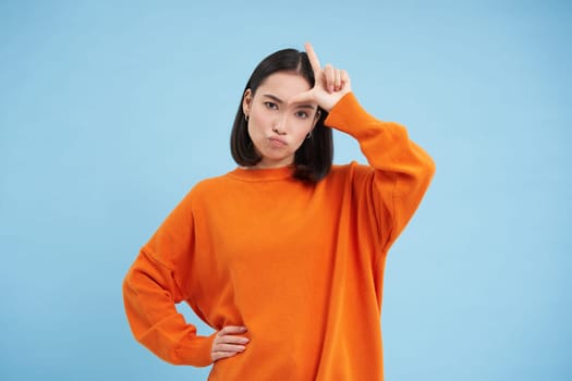 Losers. Young asian woman shows L letter on her forehead, stares sassy and confident at camera, stands over blue background.