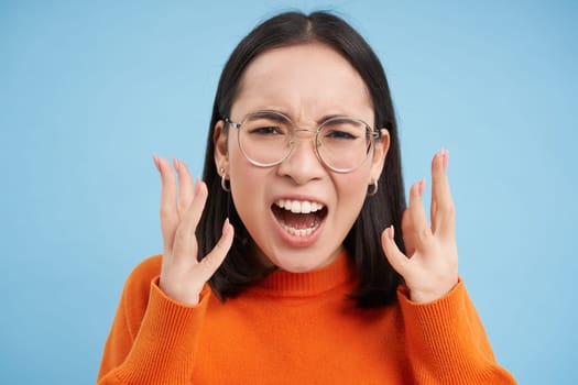 Portrait of angry japanese girl in glasses, screaming and looking furious, shaking hands in anger, looks outraged, stands over blue background.