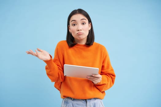 Portrait of unsure girl, asian woman with digital tablet, shrugging shoulders and looks complicated, standing over blue background.