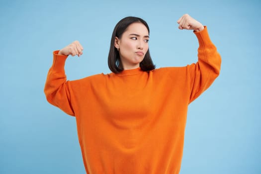 Portrait of cute korean woman flexing her biceps, shows strong arms, muscles and smiles, concept of healthy and fit people, blue background.
