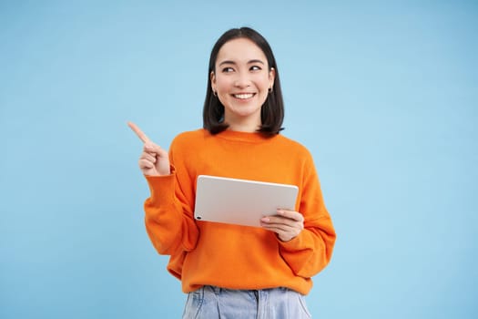 Modern asian woman with tablet, pointing at empty space for promo offer, banner, smiling and looking at advertisement, blue background.