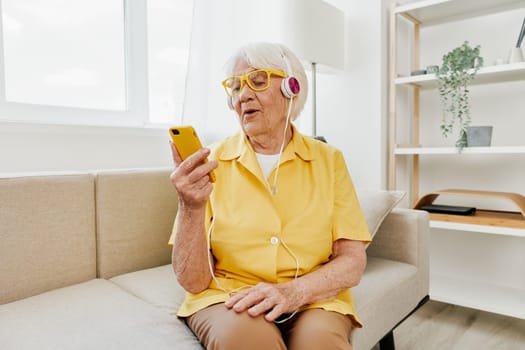 Happy elderly woman looking into her phone video call with headphones smile, technology for communication, bright modern interior, lifestyle online communication. High quality photo