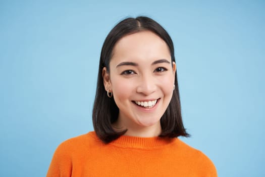 Close up portrait of beautiful korean woman with healthy smile, natural clear facial skin, stands over blue background.
