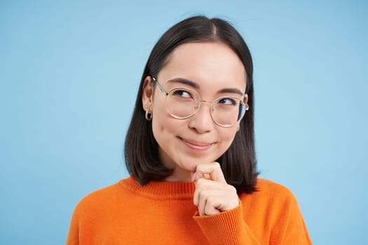 Portrait of thoughtful korean woman in glasses, thinking and looking aside, has an idea, plans smth, stands over blue background.