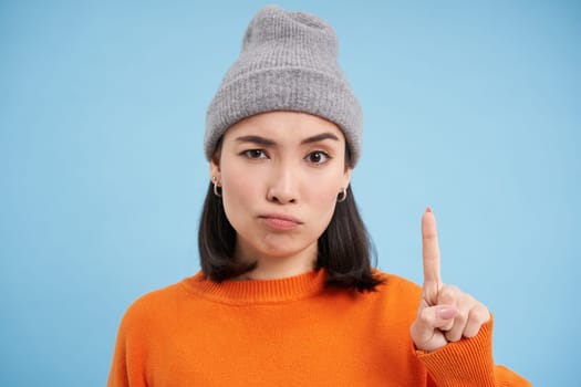 Asian girl in hat scolding you, shakes finger in disapproval, disappointed by behavious, standing over blue background.