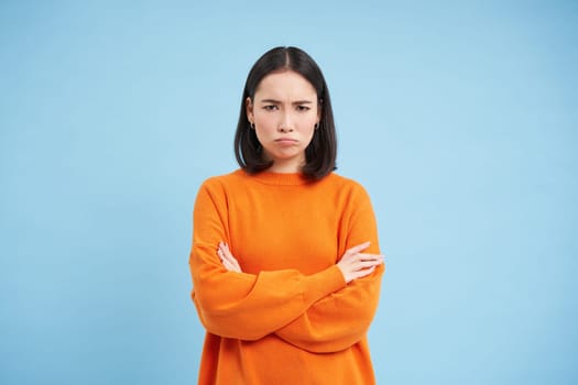 Defensive asian woman shuts herself from conversation, cross hands on chest, sulks and frowns with angry face, stands over blue background