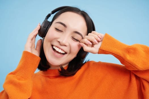 Close up of dancing asian woman in headphones, listening music in earphones and smiling happy, blue background.