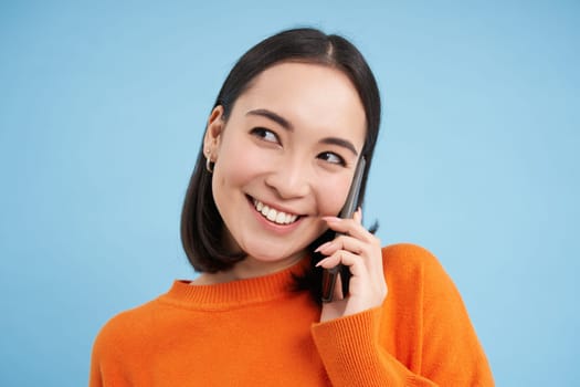 Technology concept. Close up of smiling asian woman talks on mobile phone, having conversation on cellphone, blue background.