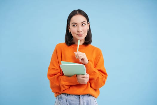 Education and students. Happy asian woman, holding notebooks and laughing, smiling at camera, enjoys going to University or College, blue background.