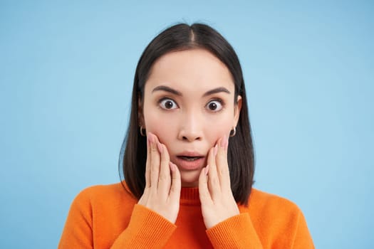 Close up portrait of asian girl with shocked face, looks at smth unbelievable, startled by news, stands over blue background.