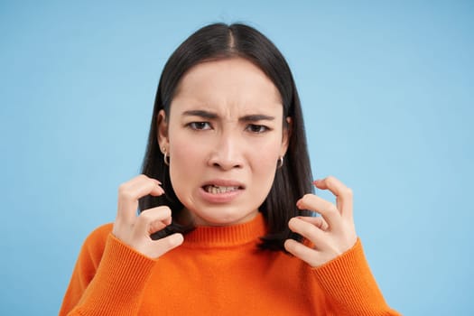 Close up of angry japanese woman, clenches her fists and looks furious, outraged and annoyed, stands over blue background.